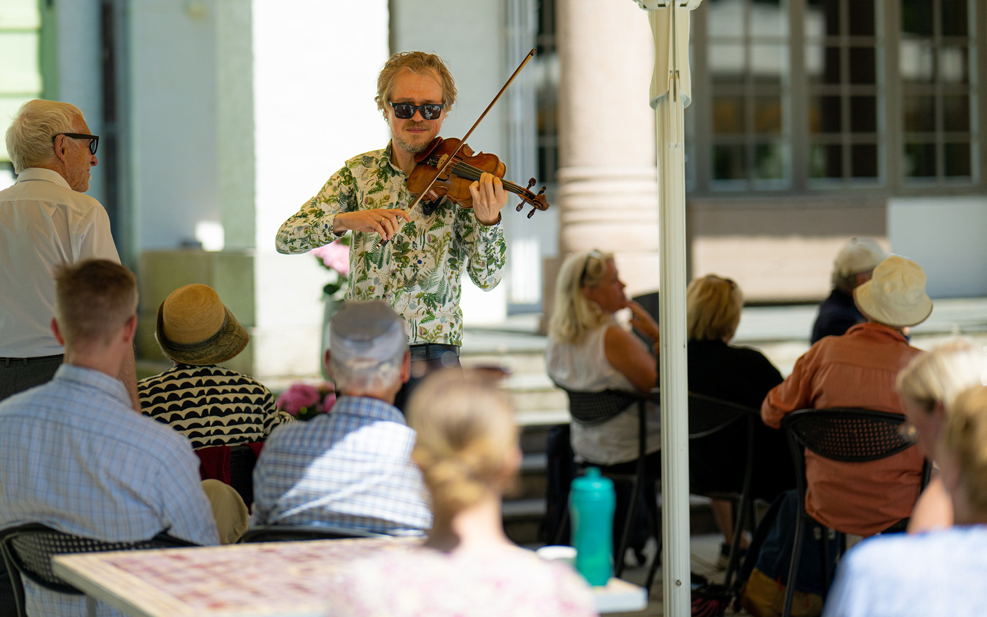 Ung mann spiller fiolin utendørs i blomstrete skjorte. Eldre publikum.