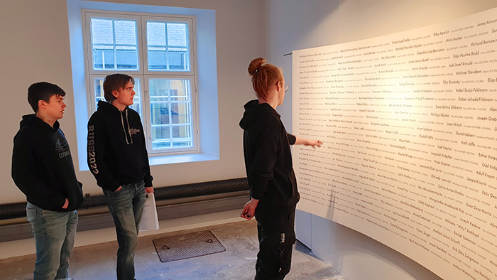 A room with names written on the wall. Three young people are standing in the room reading the names.