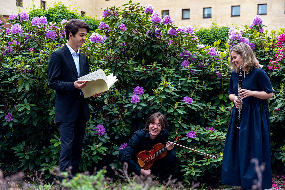 Bildet viser to unge menn og en kvinne med noter og fiolin foran en  grønn busk med lilla blomster
