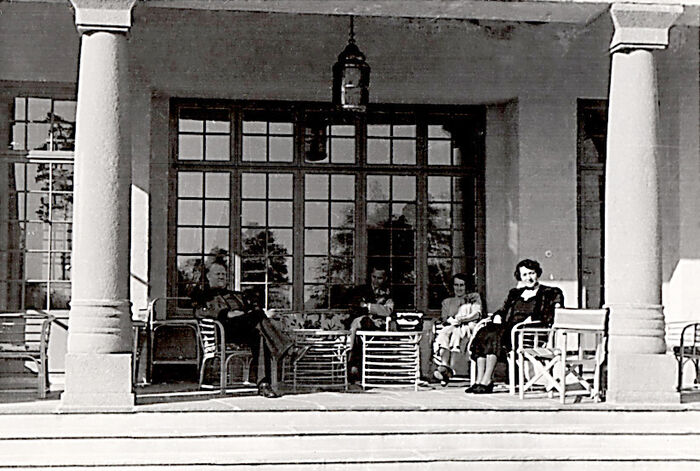 To menn og to kvinner sitter på en terasse. Den ene kvinne n har et barn på armen. Sort.hvitt foto