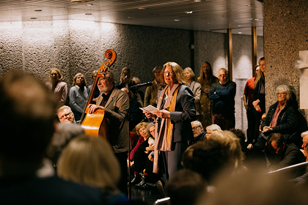 Kvinne i 60-årene med et papir i hånden foran en mikrofon, en mann med cello på venstre side, mange mennesker rundt