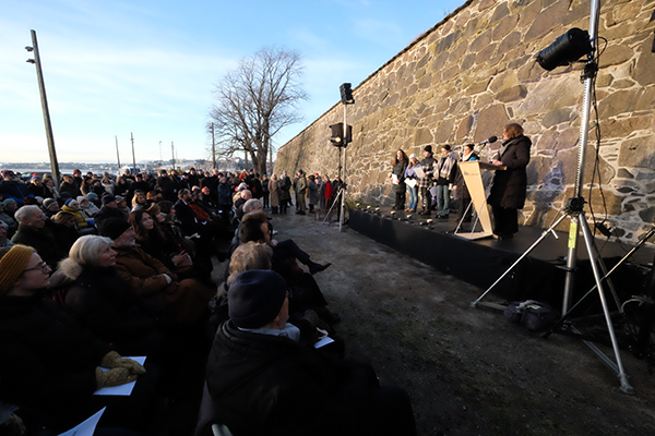 Kvinne som taler til en forsamling utendørs. Blp himmel i bakgrunnen.
