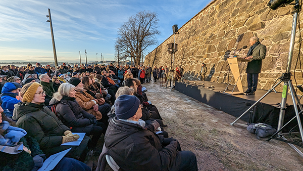 Bildet viser en forsamling som sitter utendørs og en man. som taler fra et podiet. Det er blå himmel i bakgrunnen.