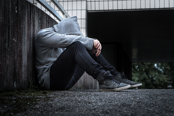 Mann sitter på bakken med hettegenser og hetten over hodet. Ser trist ut.