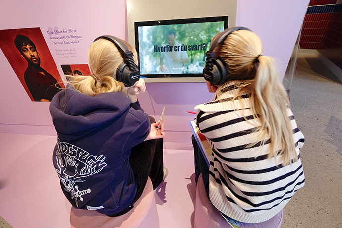 To unge jenter med hodetelefoner ser og hører på et utstillingselement i stillingen om hverdagsrasisme