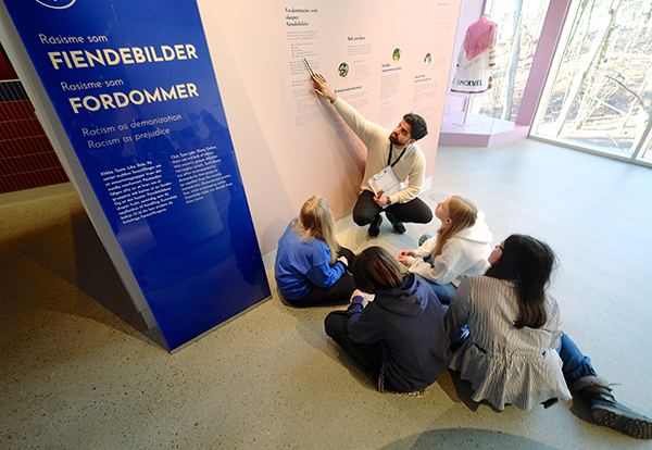 En man sitter på huk foran en vegg med teksten "Rasisme som fiendebilder, rasisme som fordommer. Foran sitter det fire barn i 10-års alderen