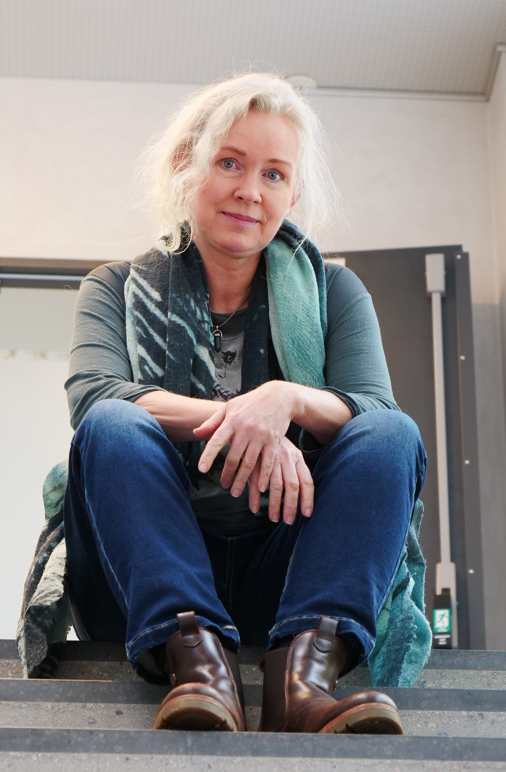 Kvnne med blondt hår, grønn genser og blå bukse sitter i en trapp. Ser i kameraet med et alvorlig uttrykk.