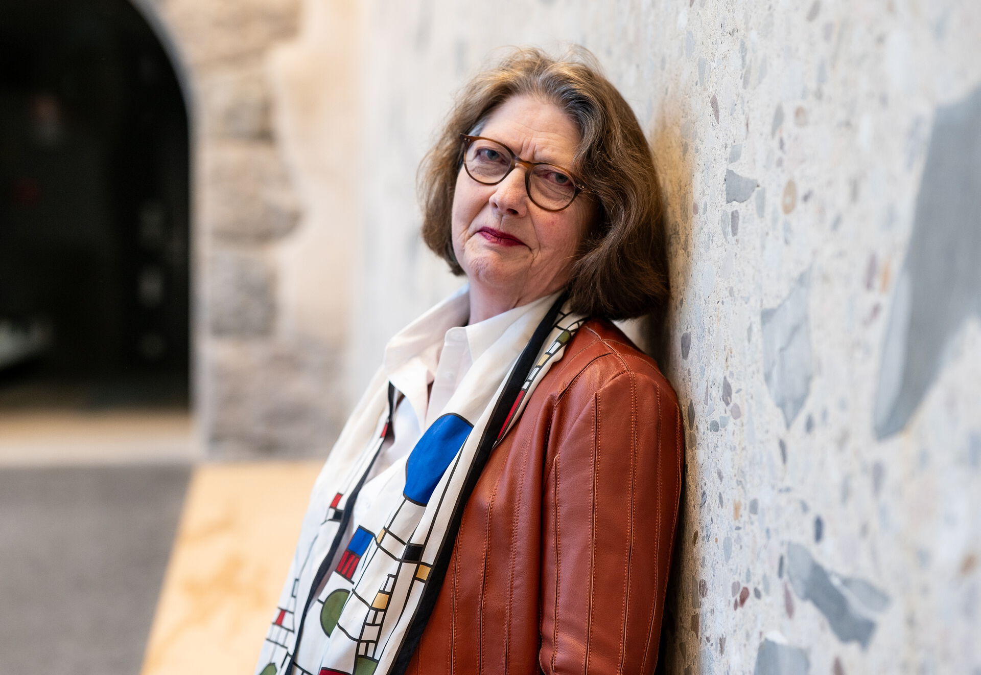Voksen dame med halvlangt hår, skinnjakke og et skjerf i halsen, som lener seg mot en murvegg