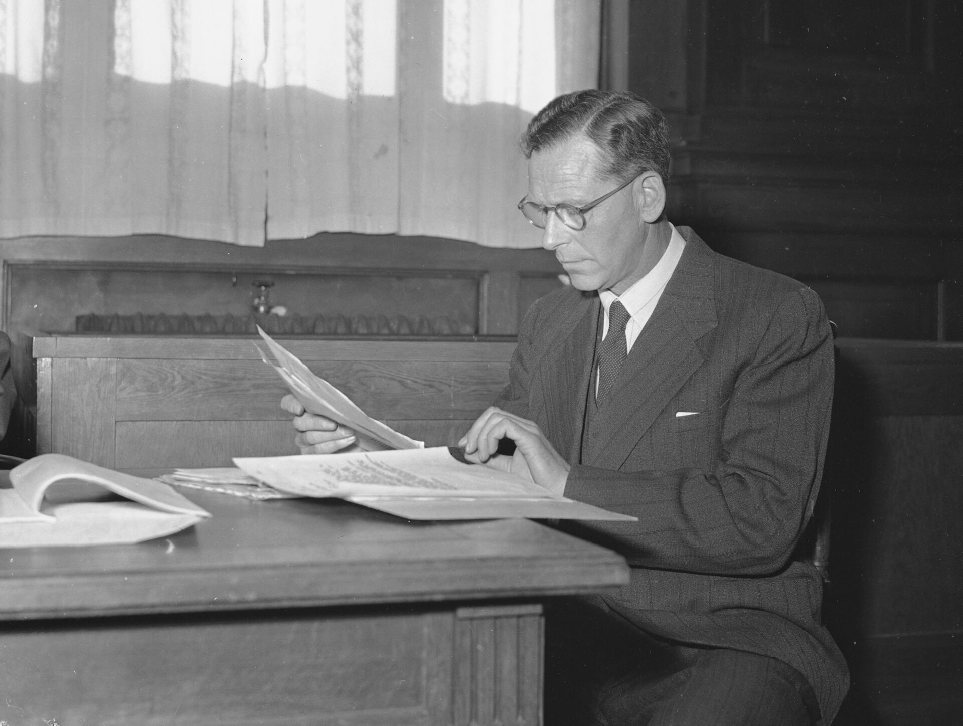 Svart/hvitt-bilde av en voksen mann i dress som sitter ved et bodr og blar i noen papirer
