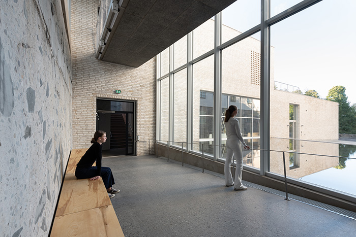 A young girl are sitting on a bench, looking out the window. Another girl stands in the room, also looking out the window